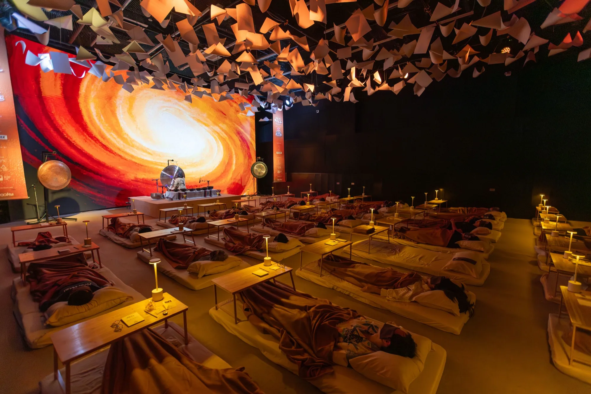 A full sound bath event in progress, guests resting on mats before a lit stage and gong.